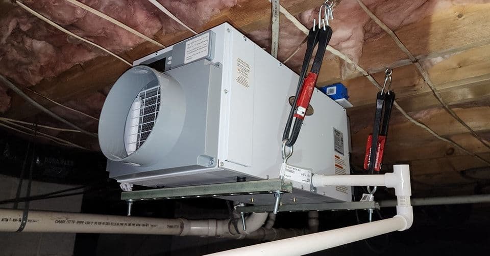Basement ventilation system mounted on ceiling with piping and insulation in background.