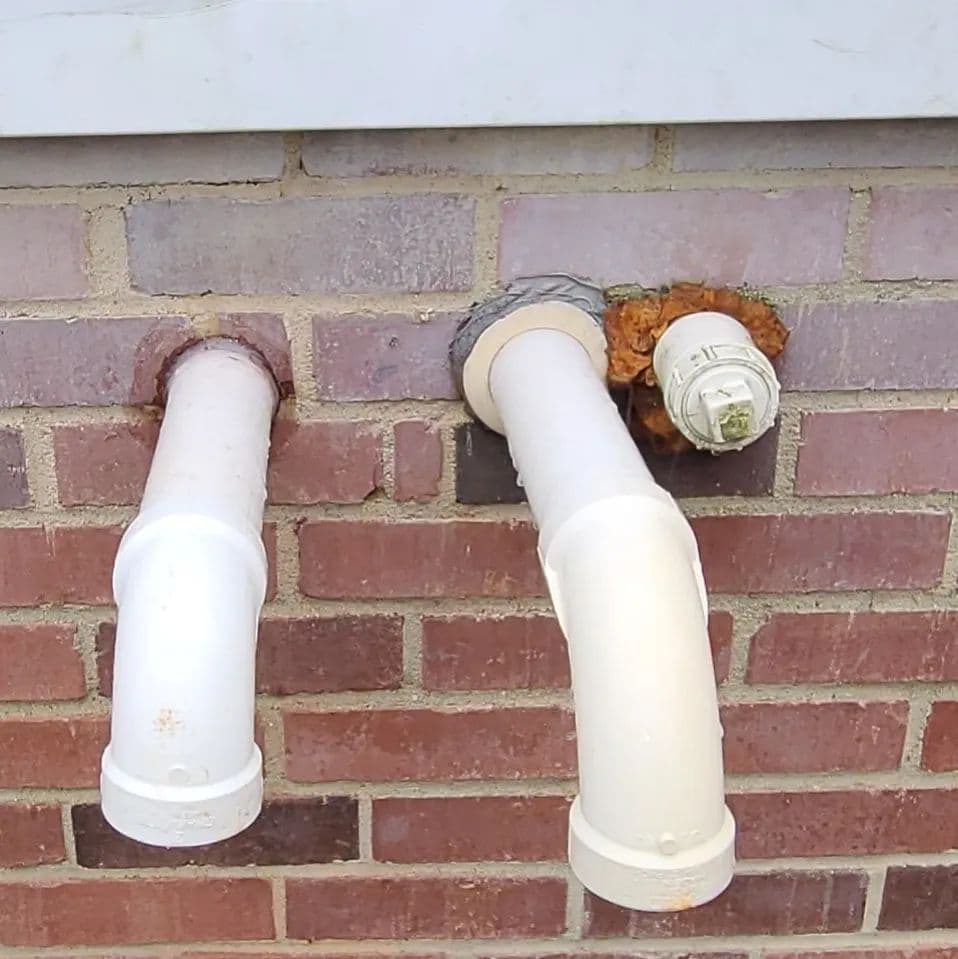 Two white PVC pipes on a brick wall, one showing signs of corrosion.