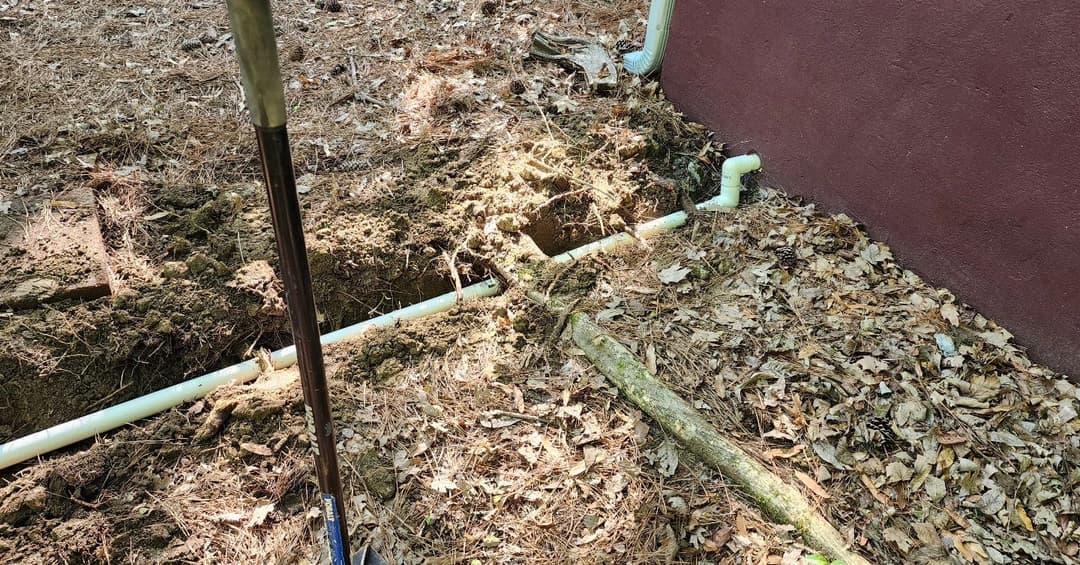 Excavated area showing newly laid white PVC pipes near building foundation in wooded setting.