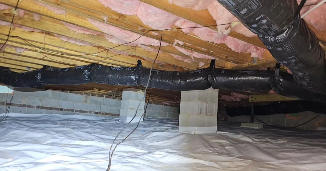 View of a crawl space with insulation, black ductwork, and a vapor barrier covering the ground.