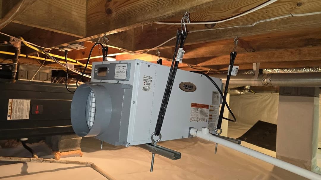 Air exchanger unit installed in a basement, suspended from wooden beams with ventilation pipes.