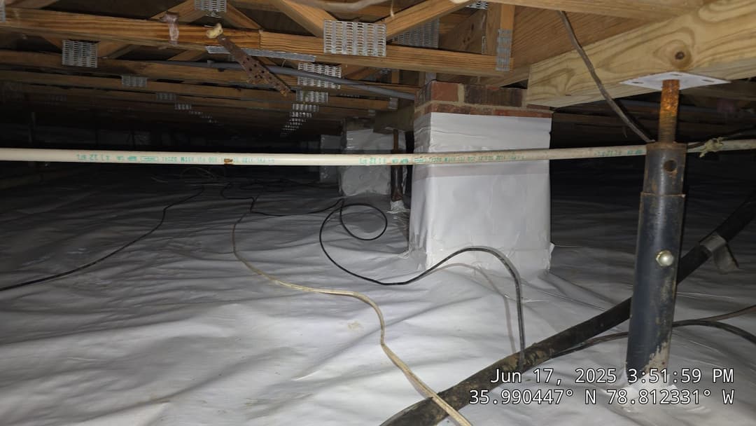 Underhouse foundation with support columns and plastic sheeting, showcasing plumbing and wiring.