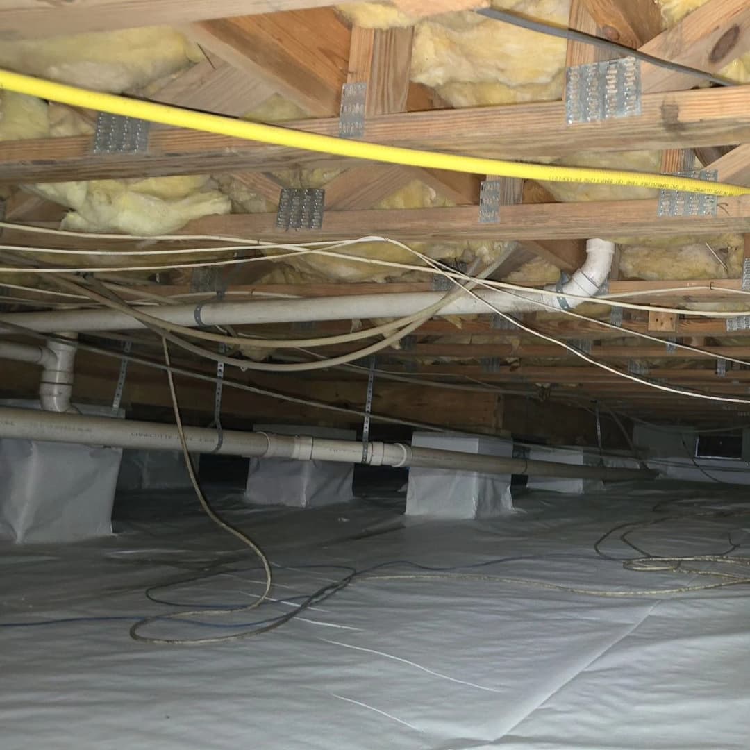View of a crawl space with insulation, pipes, and wiring, illustrating home maintenance concerns.