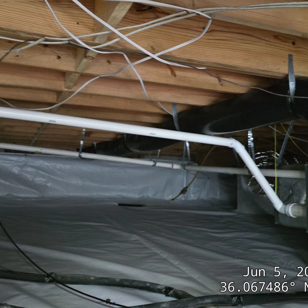 Exposed basement plumbing and electrical wiring under wooden beams with white vapor barrier.