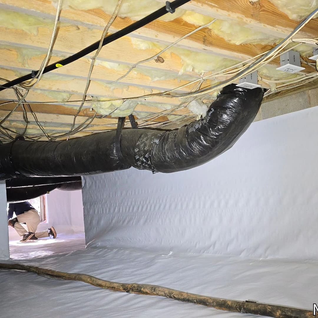 Crawl space with black ductwork, insulation, and a worker inspecting the area.