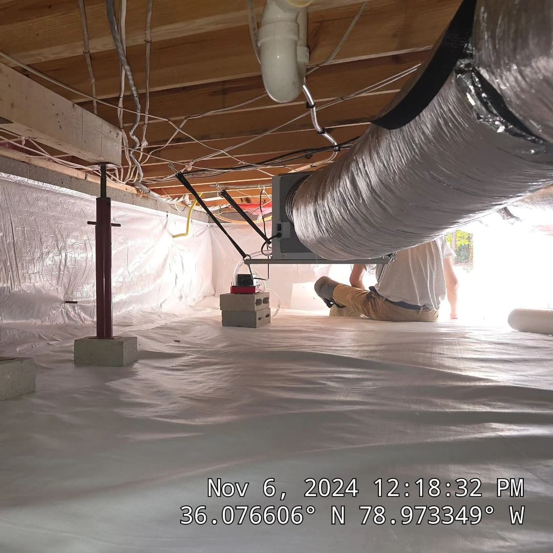 Person working under a house with insulation and ductwork, showcasing crawl space maintenance.
