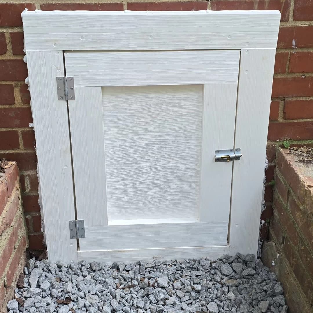 White wooden access door with silver hinges and latch, set into brick wall and gravel.