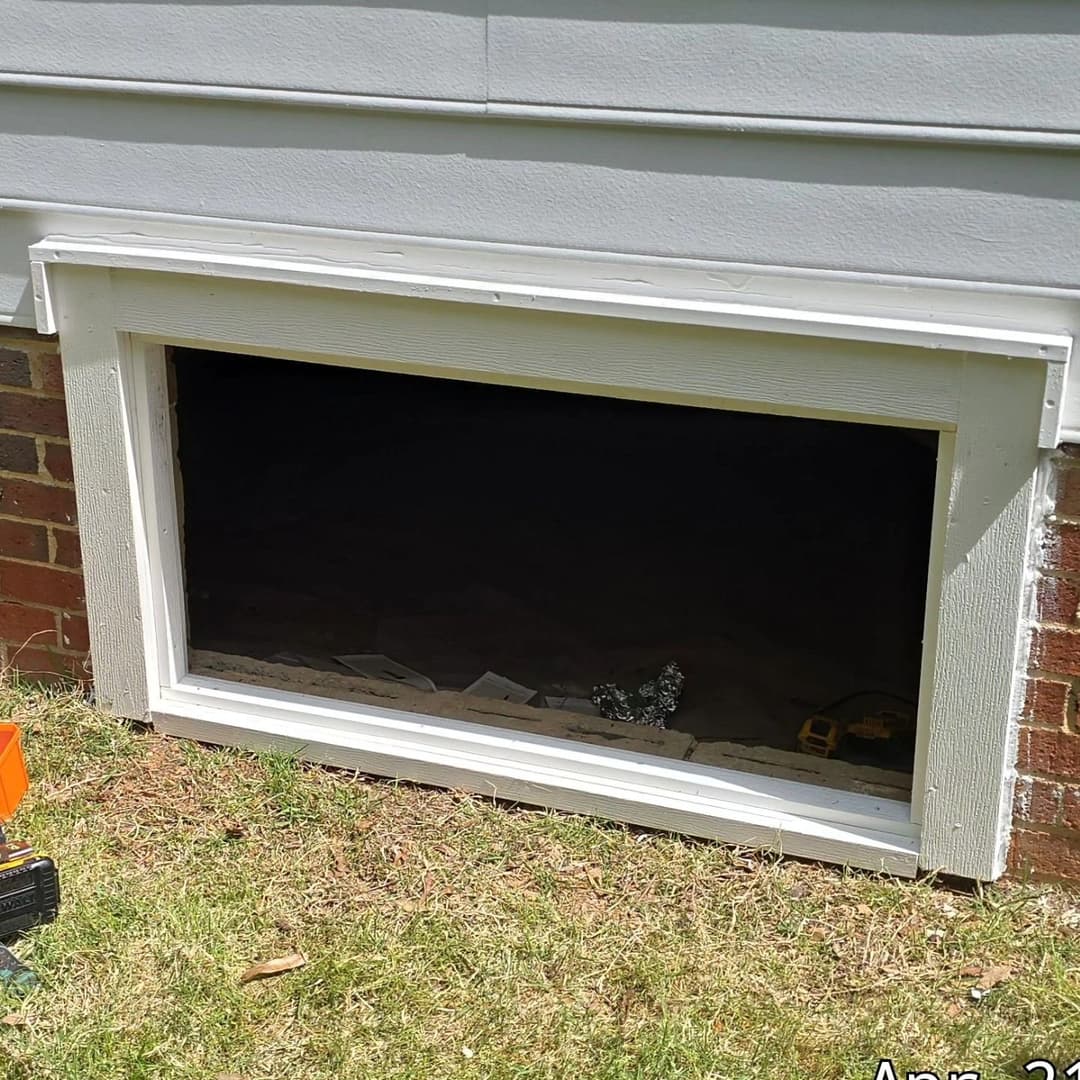 Window opening with a white frame, revealing a dark interior, surrounded by grass and tools.