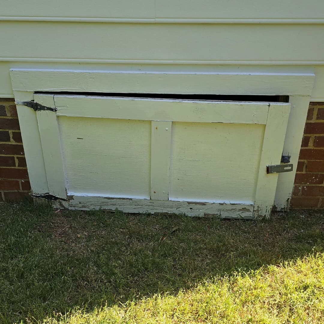 White wooden crawl space door with rusted hinges, set against a brick foundation.