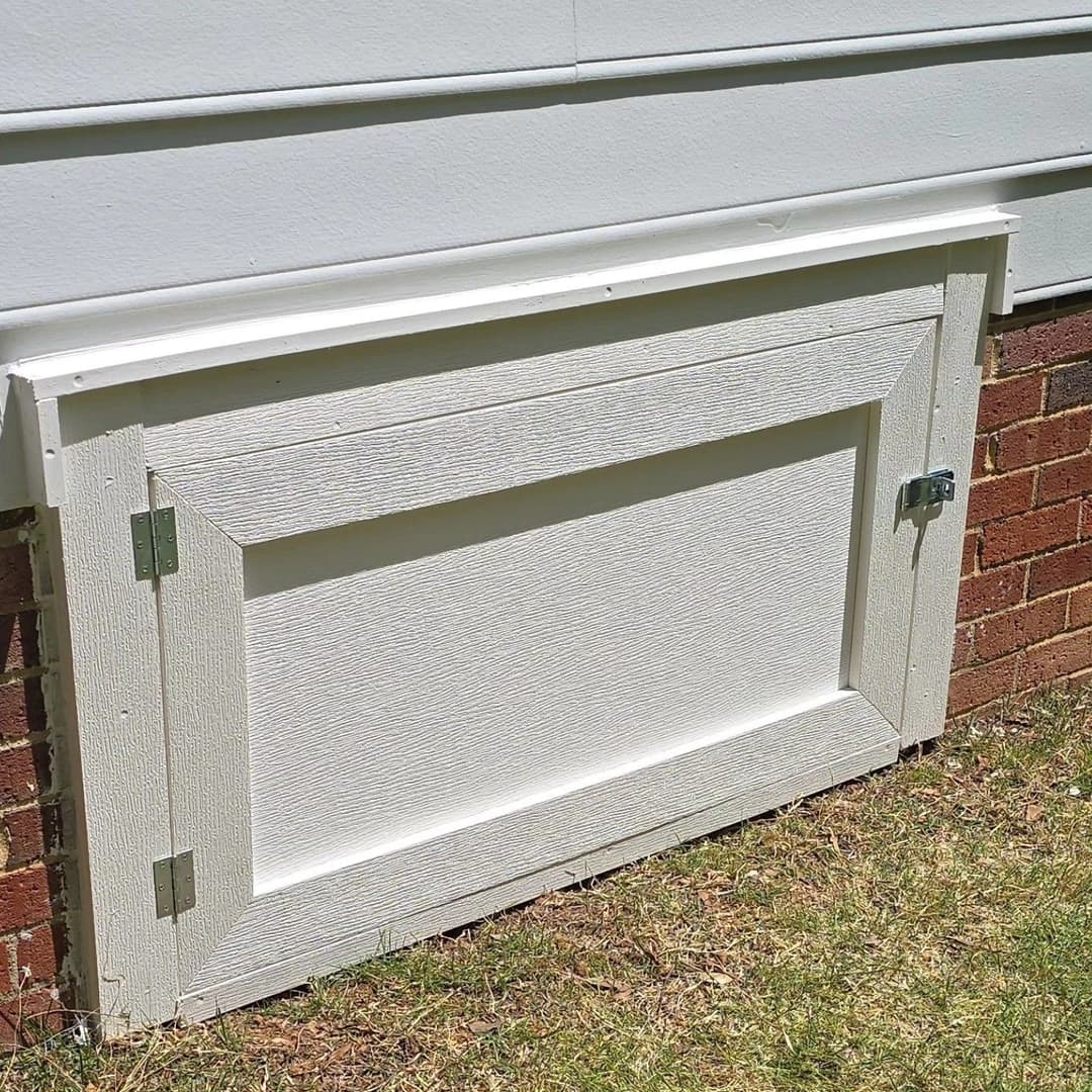 White exterior vent cover with metal latch on brick wall, surrounded by green grass.