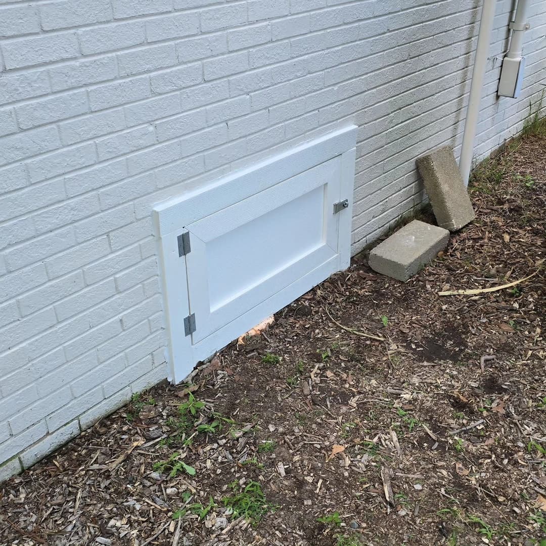 White access door on a brick wall beside landscaping, featuring metal latches and gravel.