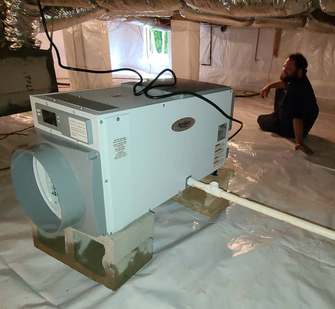 Crawl space air purifier installed on blocks with technician seated nearby in plastic-lined area.